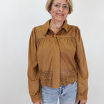 A woman modeling a brown long-sleeve blouse with a gathered yoke and eyelet lace hem, paired with light blue jeans against a plain white background.