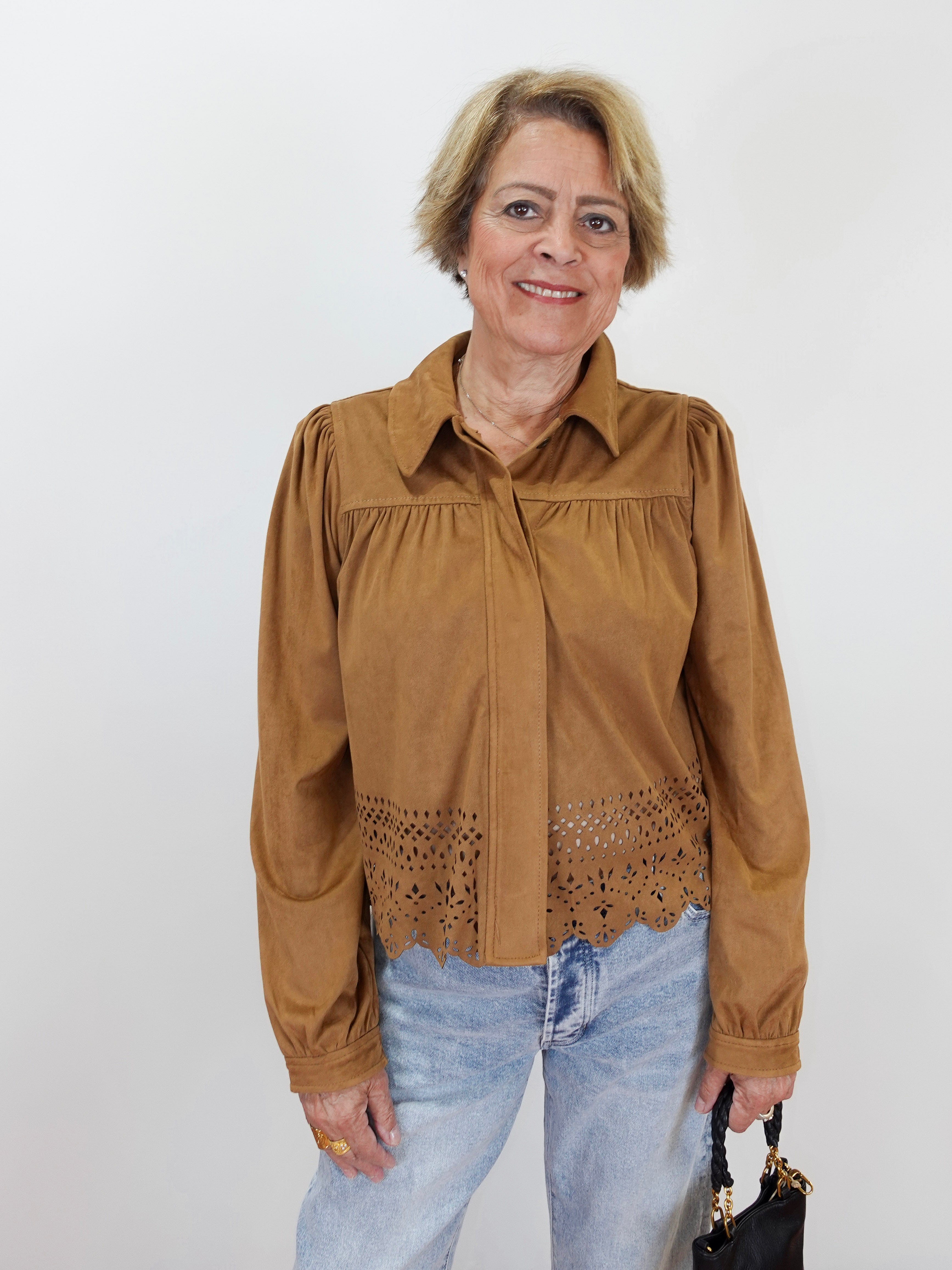 A woman modeling a brown long-sleeve blouse with a gathered yoke and eyelet lace hem, paired with light blue jeans against a plain white background.