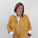 A woman wearing a mustard yellow utility jacket over a white collared shirt and dark pants, standing against a plain light gray background.