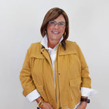 A woman wearing a mustard yellow utility jacket over a white collared shirt and dark pants, standing against a plain light gray background.