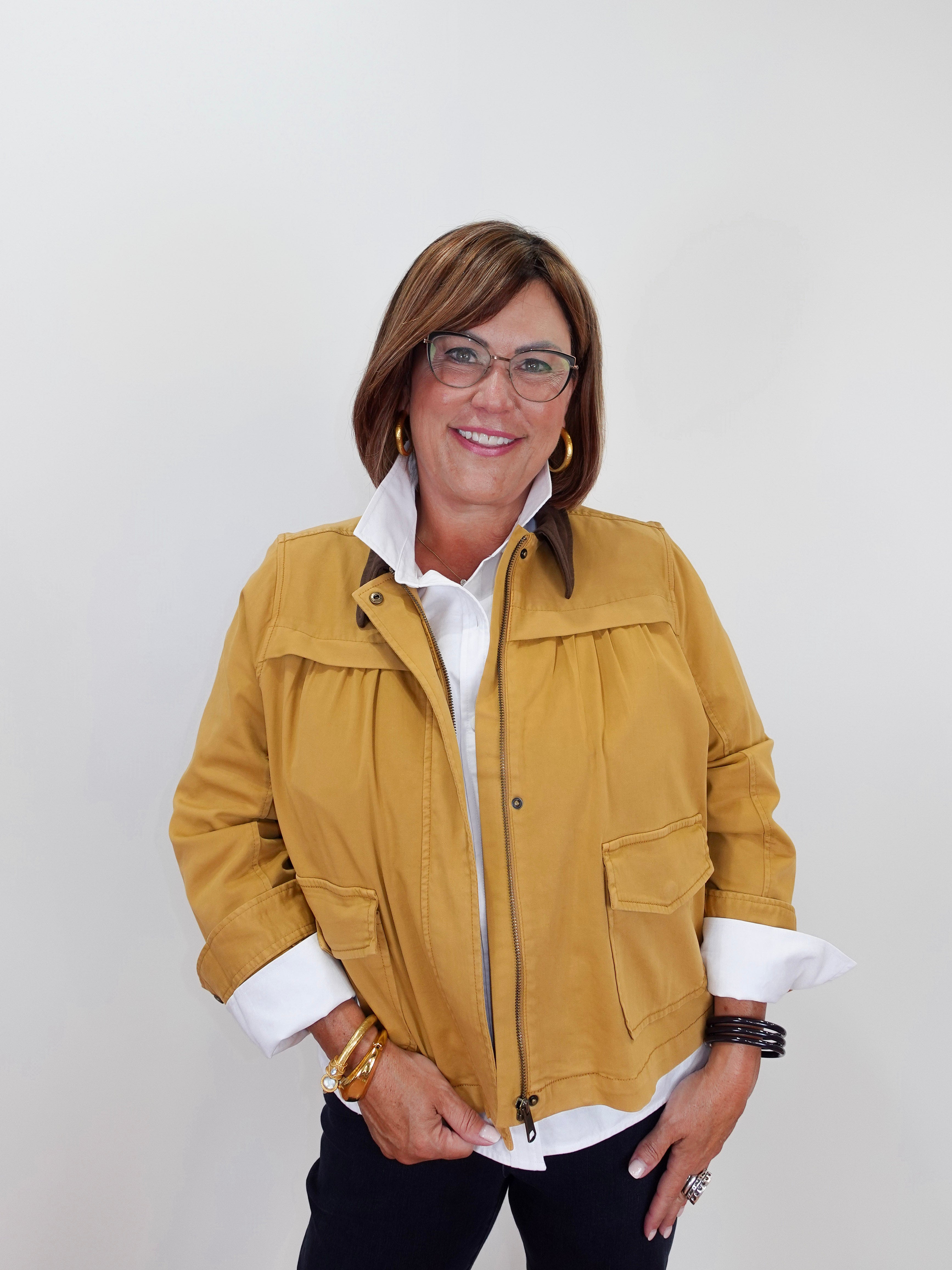 A woman wearing a mustard yellow utility jacket over a white collared shirt and dark pants, standing against a plain light gray background.