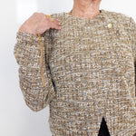 A close-up of a woman wearing a textured tweed jacket in beige, brown, and white with gold-toned zipper and button details, paired with dark brown pants.