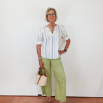 Woman wearing a striped shirt and green pants standing against a white wall.