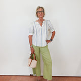 Woman wearing a striped shirt and green pants standing against a white wall.