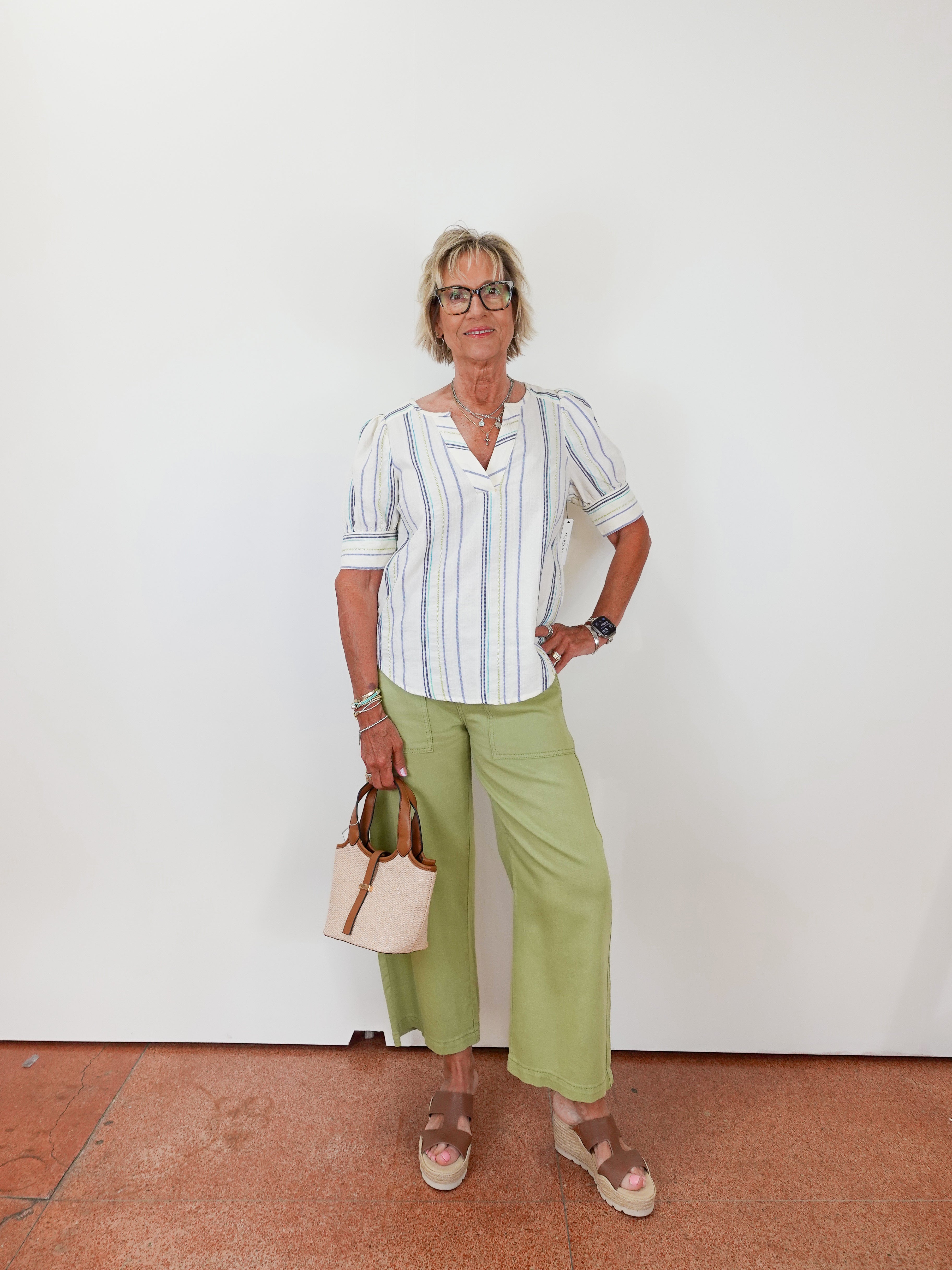 Woman wearing a striped shirt and green pants standing against a white wall.