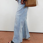 A person wearing light-wash wide-leg denim jeans and black slide sandals, carrying a tan crossbody bag against a plain white wall.