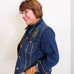 Woman wearing a blue denim jacket with a brooch, standing against a white wall.