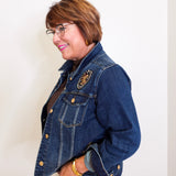 Woman wearing a blue denim jacket with a brooch, standing against a white wall.