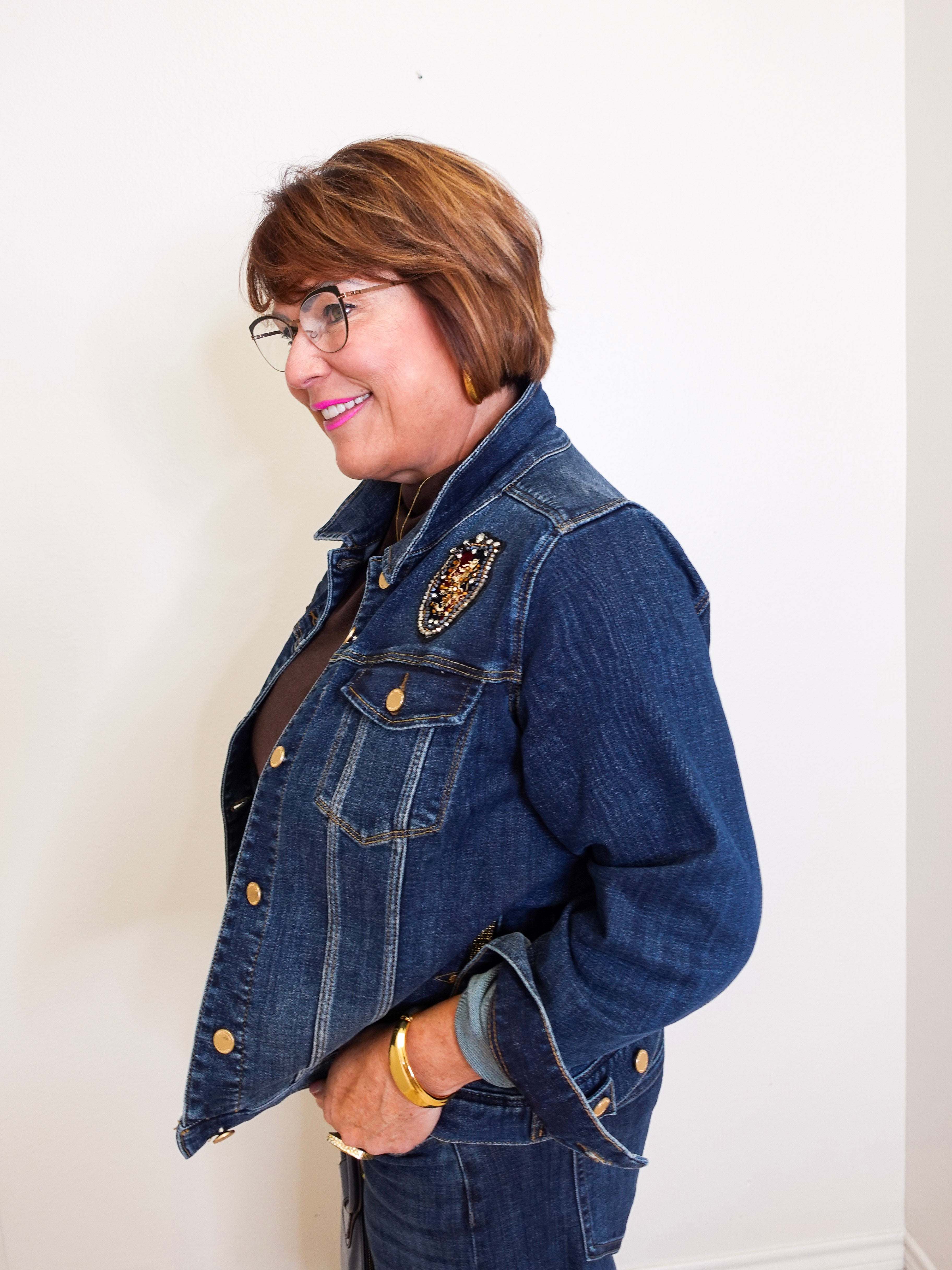 Woman wearing a blue denim jacket with a brooch, standing against a white wall.