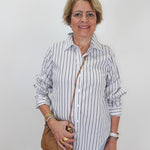 A smiling older woman with short blonde hair stands against a plain light gray background, wearing a white button-up shirt with thin dark vertical stripes, light blue jeans, and holding a small tan crossbody bag.