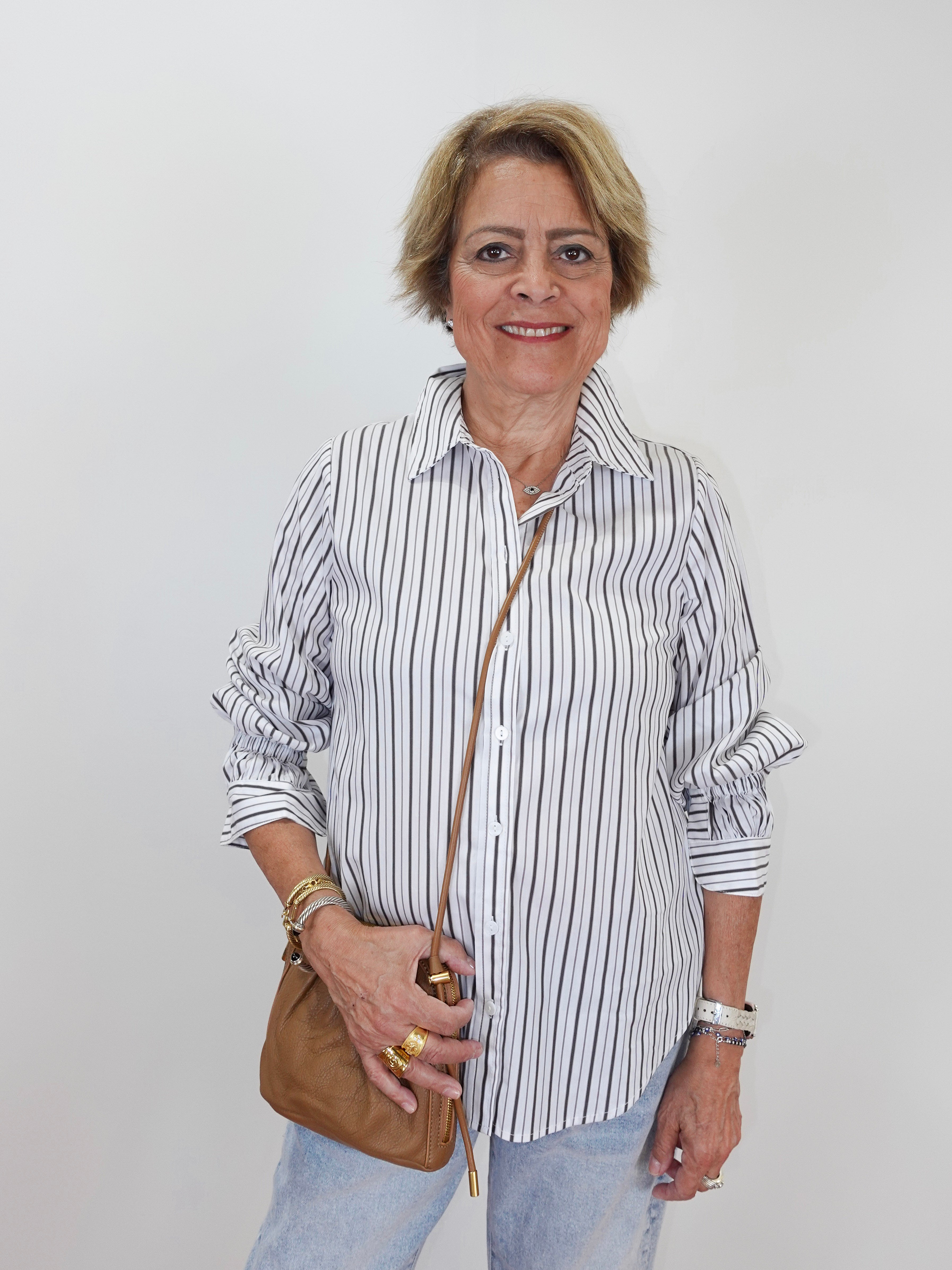 A smiling older woman with short blonde hair stands against a plain light gray background, wearing a white button-up shirt with thin dark vertical stripes, light blue jeans, and holding a small tan crossbody bag.