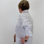 A woman shown from the side profile wearing a white button-up shirt with thin black vertical stripes, paired with light blue jeans, standing against a plain light gray wall.