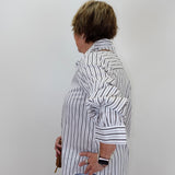 A woman shown from the side profile wearing a white button-up shirt with thin black vertical stripes, paired with light blue jeans, standing against a plain light gray wall.