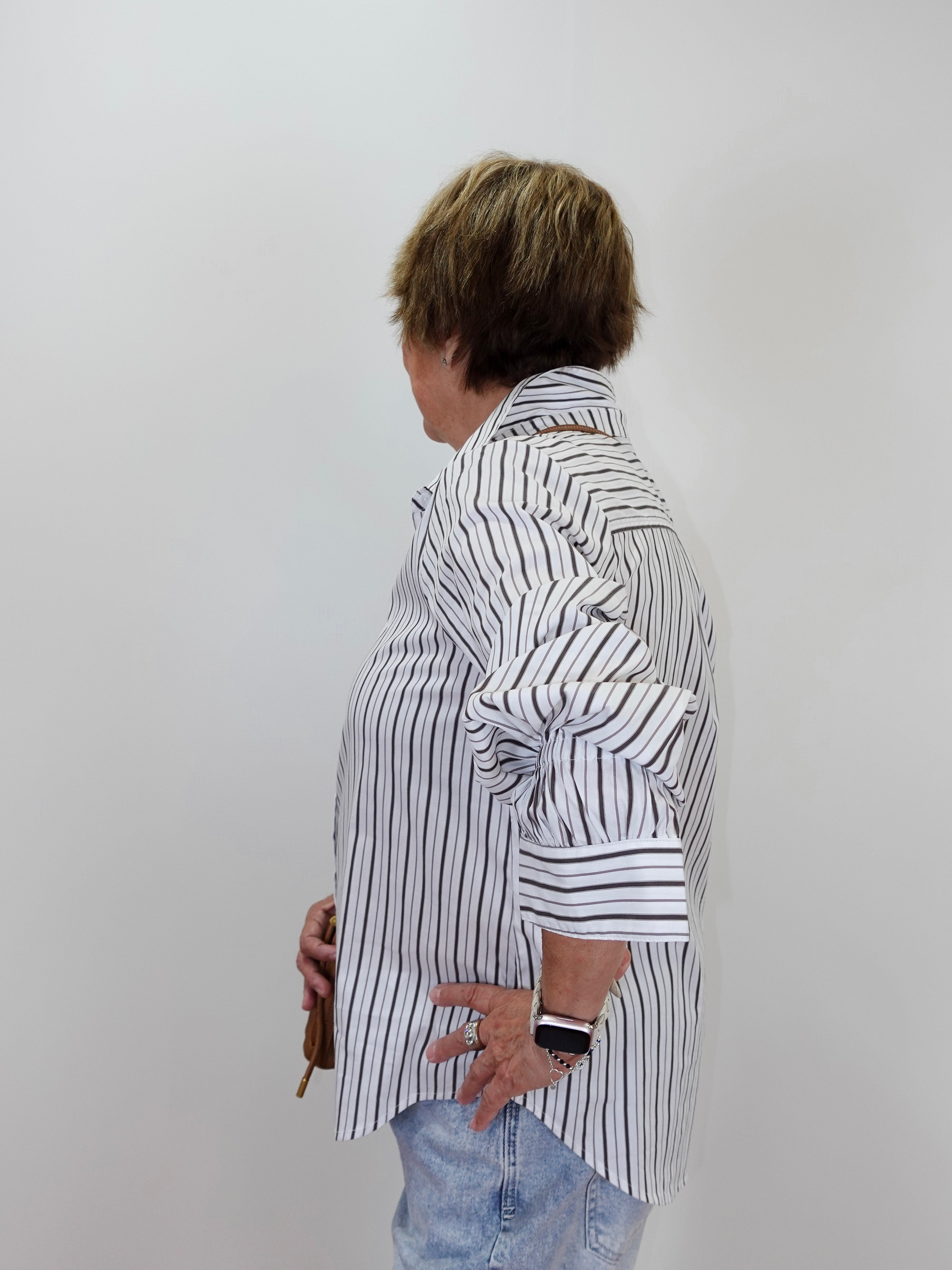 A woman shown from the side profile wearing a white button-up shirt with thin black vertical stripes, paired with light blue jeans, standing against a plain light gray wall.