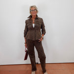 A woman wearing a matching brown patterned jacket and trousers set, holding a dark red handbag, standing against a plain white wall on a terracotta tiled floor.