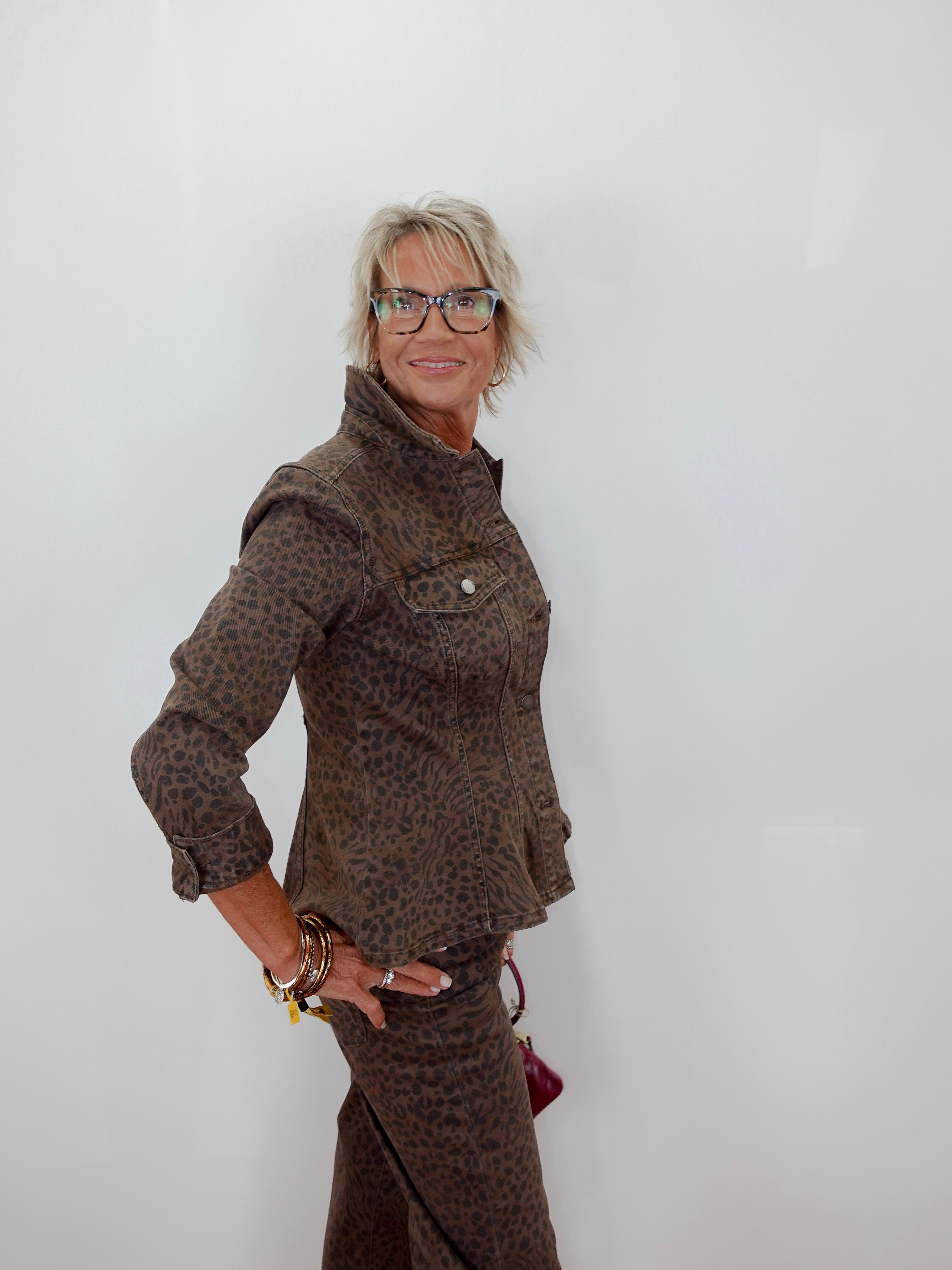 A woman with short gray hair and glasses wearing a brown leopard-print jacket and matching skirt, posing against a plain white wall.