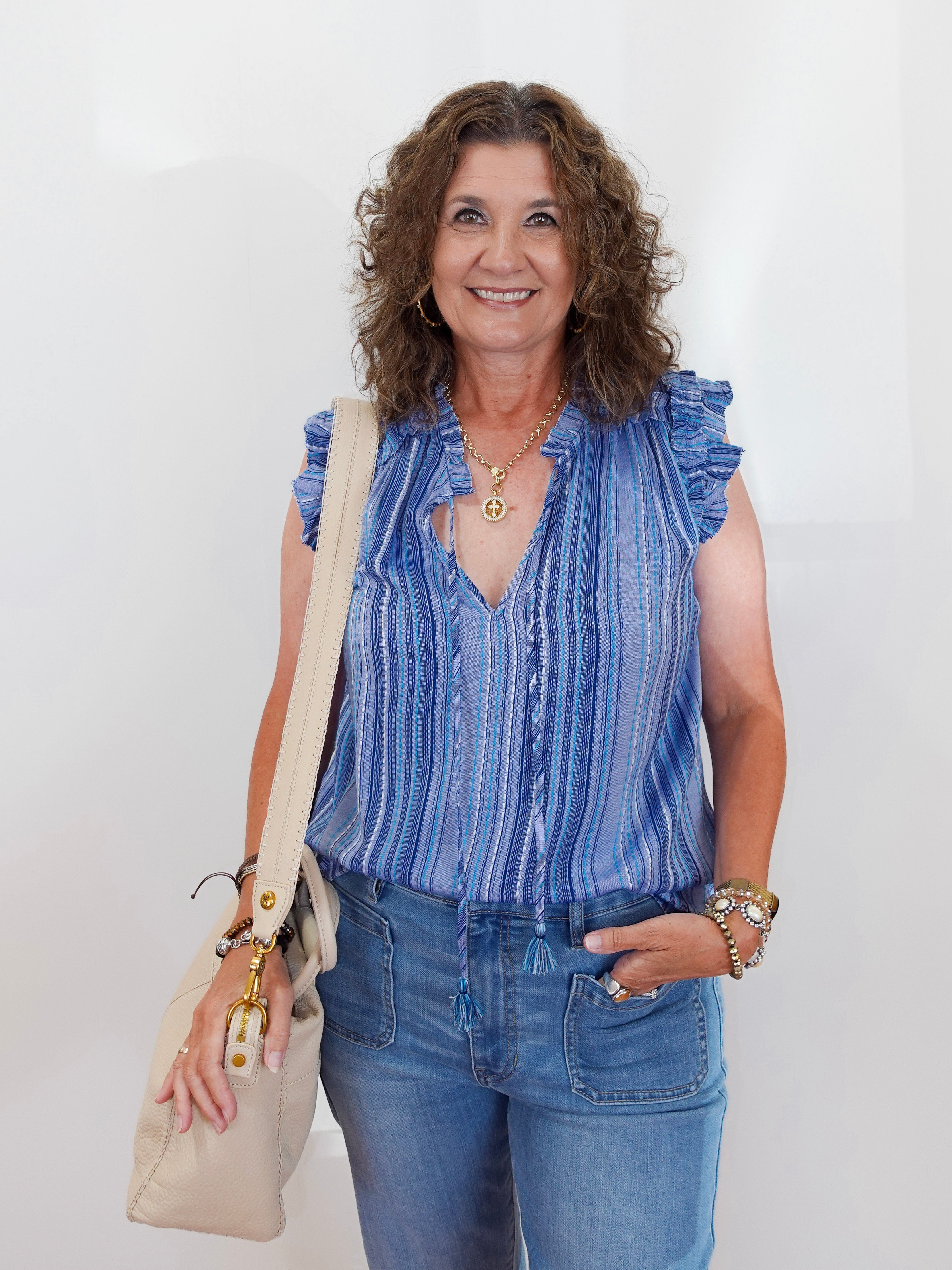 Woman wearing a blue striped sleeveless top and jeans, holding a beige bag against a white background
