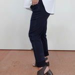 A person wearing dark navy cropped jeans with rolled cuffs, a loose white top, and dark woven heeled mule sandals, standing against a plain off-white wall and terracotta floor.