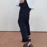 A person wearing dark navy cropped jeans with rolled cuffs, a loose white top, and dark woven heeled mule sandals, standing against a plain off-white wall and terracotta floor.
