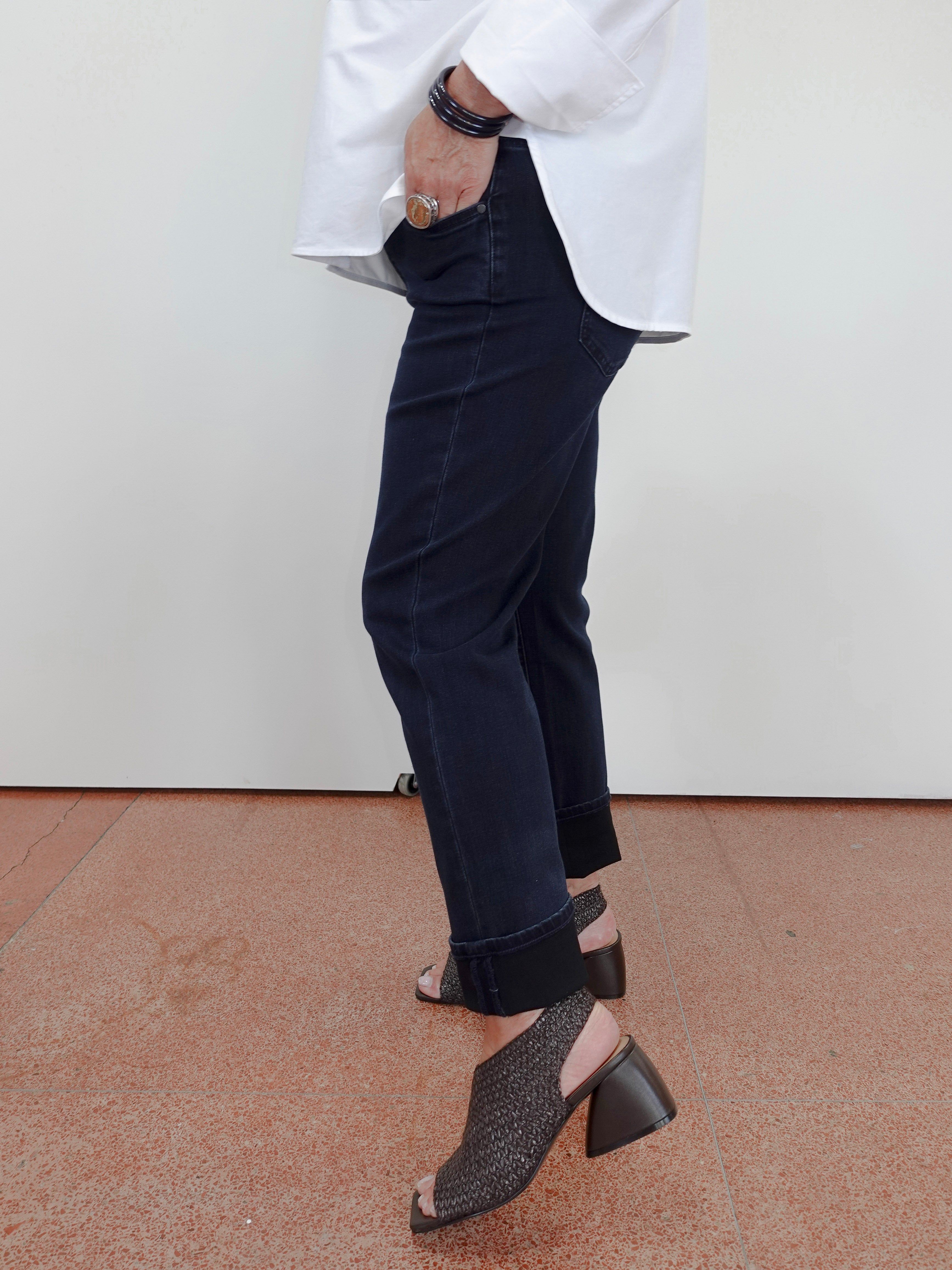 A person wearing dark navy cropped jeans with rolled cuffs, a loose white top, and dark woven heeled mule sandals, standing against a plain off-white wall and terracotta floor.