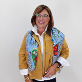 A woman wearing a mustard yellow jacket with colorful embroidered details on the front, a white collared shirt, and black pants, standing against a plain light gray background.