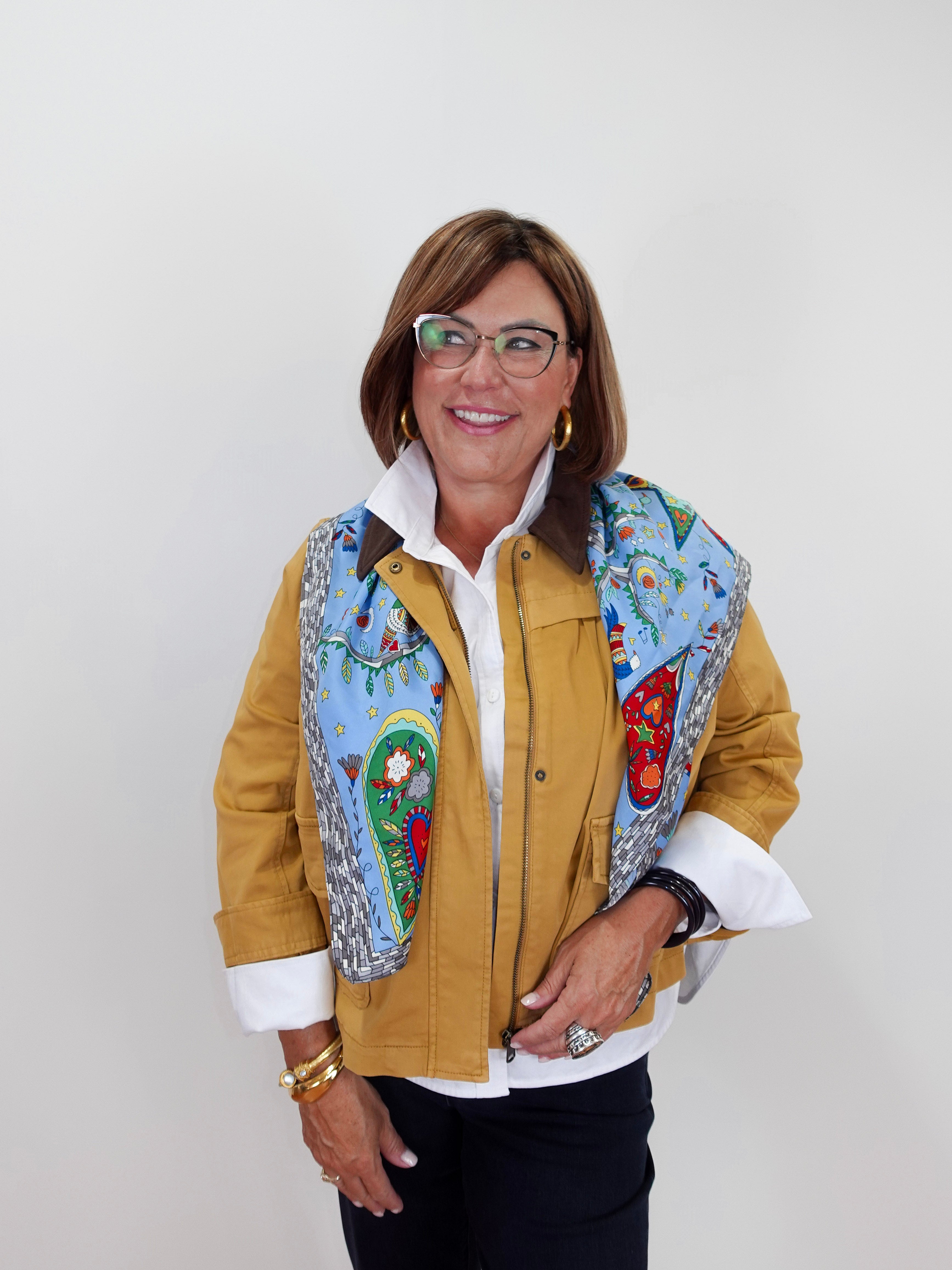 A woman wearing a mustard yellow jacket with colorful embroidered details on the front, a white collared shirt, and black pants, standing against a plain light gray background.
