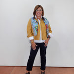 A woman wearing a mustard yellow jacket with a colorful patterned collar, a white shirt, black pants, and gray heeled sandals, standing against a plain white wall.