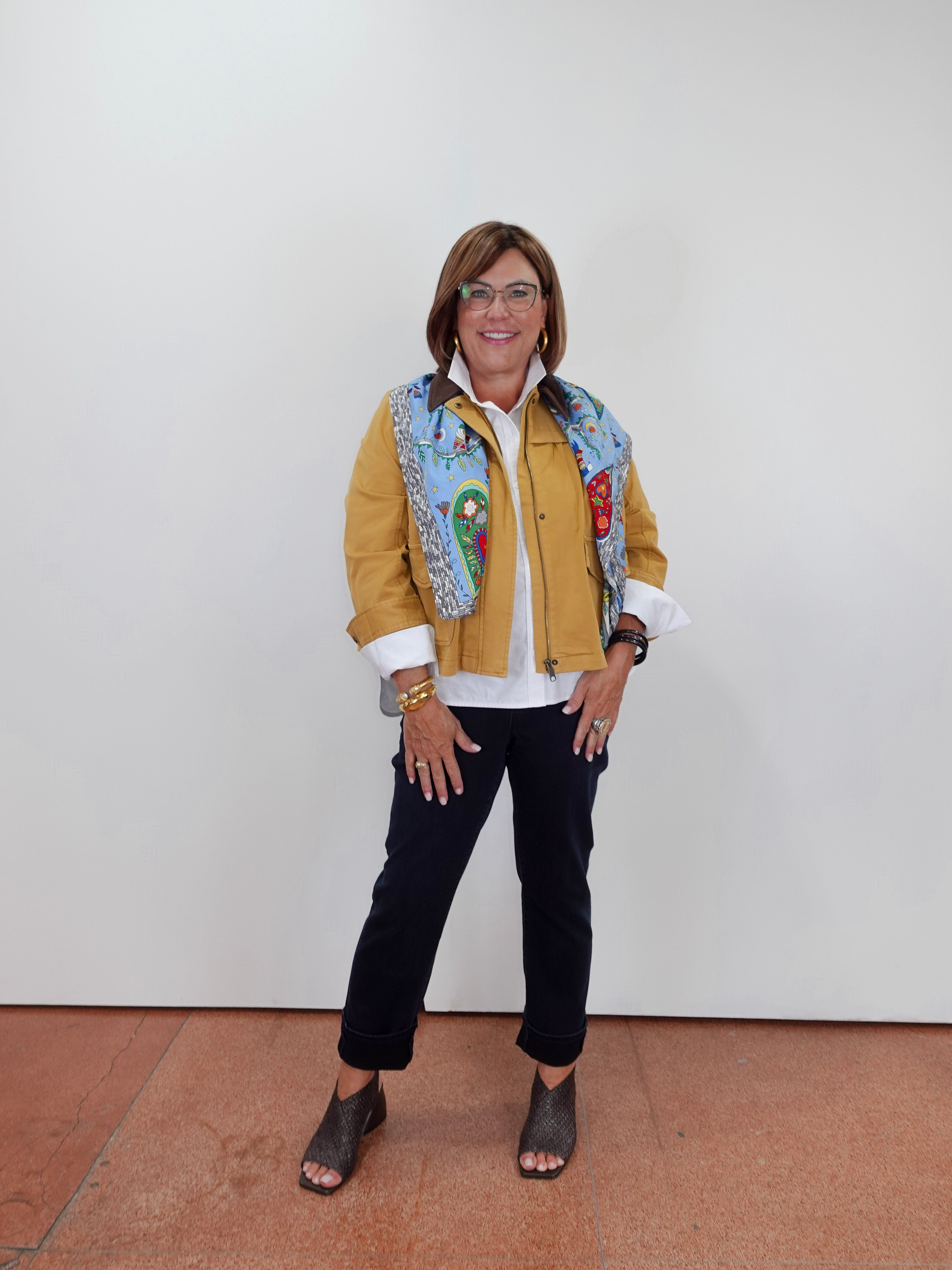A woman wearing a mustard yellow jacket with a colorful patterned collar, a white shirt, black pants, and gray heeled sandals, standing against a plain white wall.
