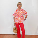 Woman wearing a colorful floral blouse and red pants, standing against a white wall.