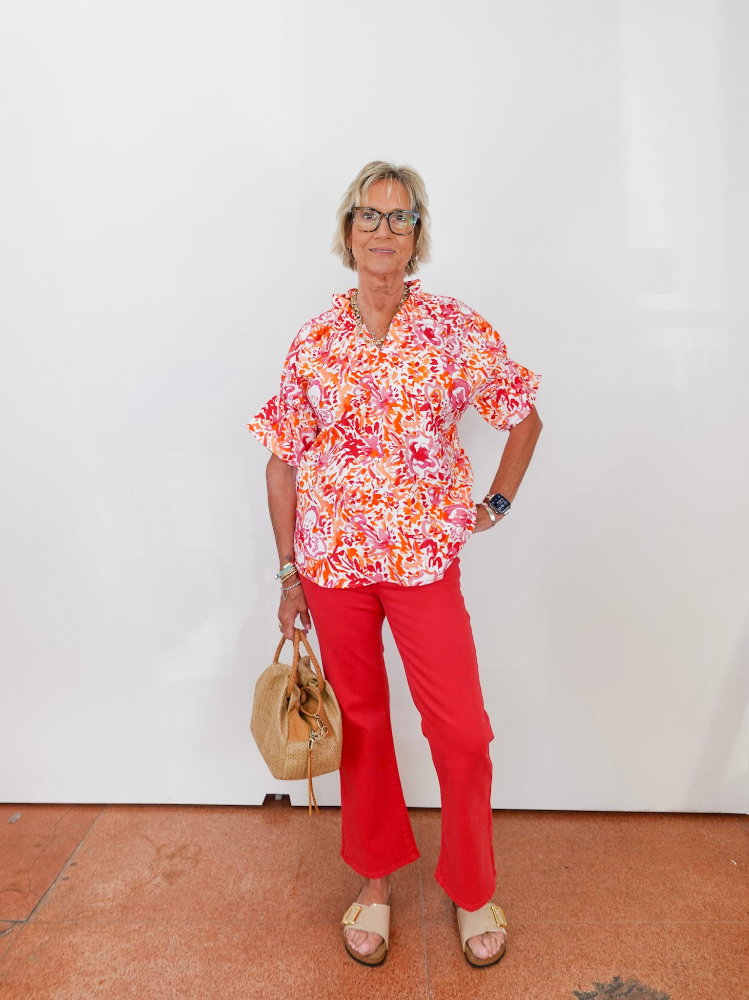 Woman wearing a colorful floral blouse and red pants, standing against a white wall.