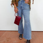 A person wearing light-wash blue flare jeans with silver stud detailing down the side, paired with a patterned blouse and black snakeskin heeled sandals, holding a small burgundy handbag against a white wall and terracotta floor.