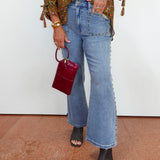 A person wearing light-wash blue flare jeans with silver stud detailing down the side, paired with a patterned blouse and black snakeskin heeled sandals, holding a small burgundy handbag against a white wall and terracotta floor.