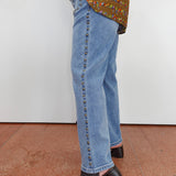 A person wearing light blue denim jeans with a vertical row of dark studs down the side, paired with a patterned top and black heeled mule sandals, standing against a plain white wall.