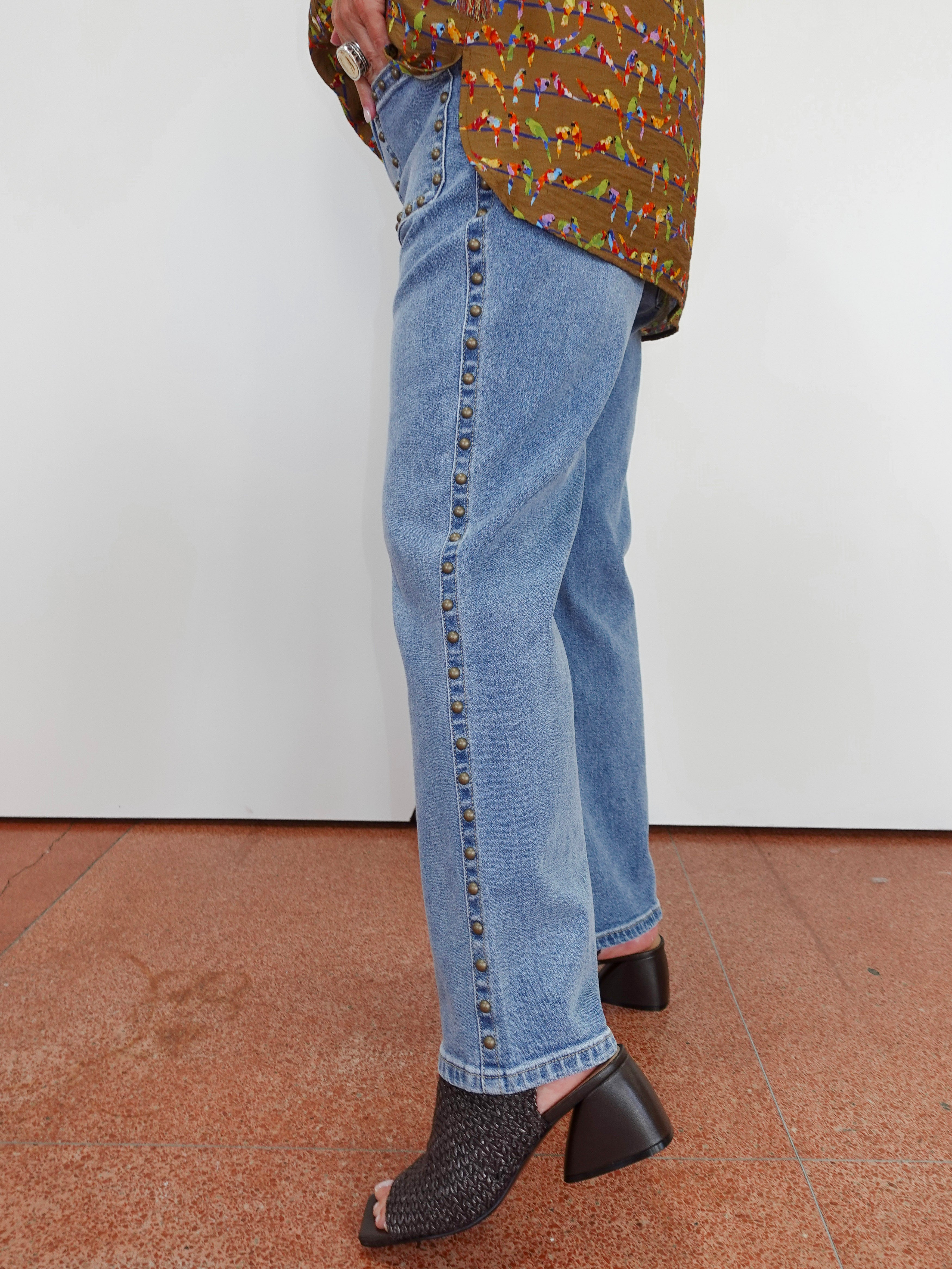 A person wearing light blue denim jeans with a vertical row of dark studs down the side, paired with a patterned top and black heeled mule sandals, standing against a plain white wall.