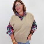 A woman wearing a beige knit V-neck sweater vest over a colorful patterned collared shirt and blue jeans, standing against a plain light gray background.