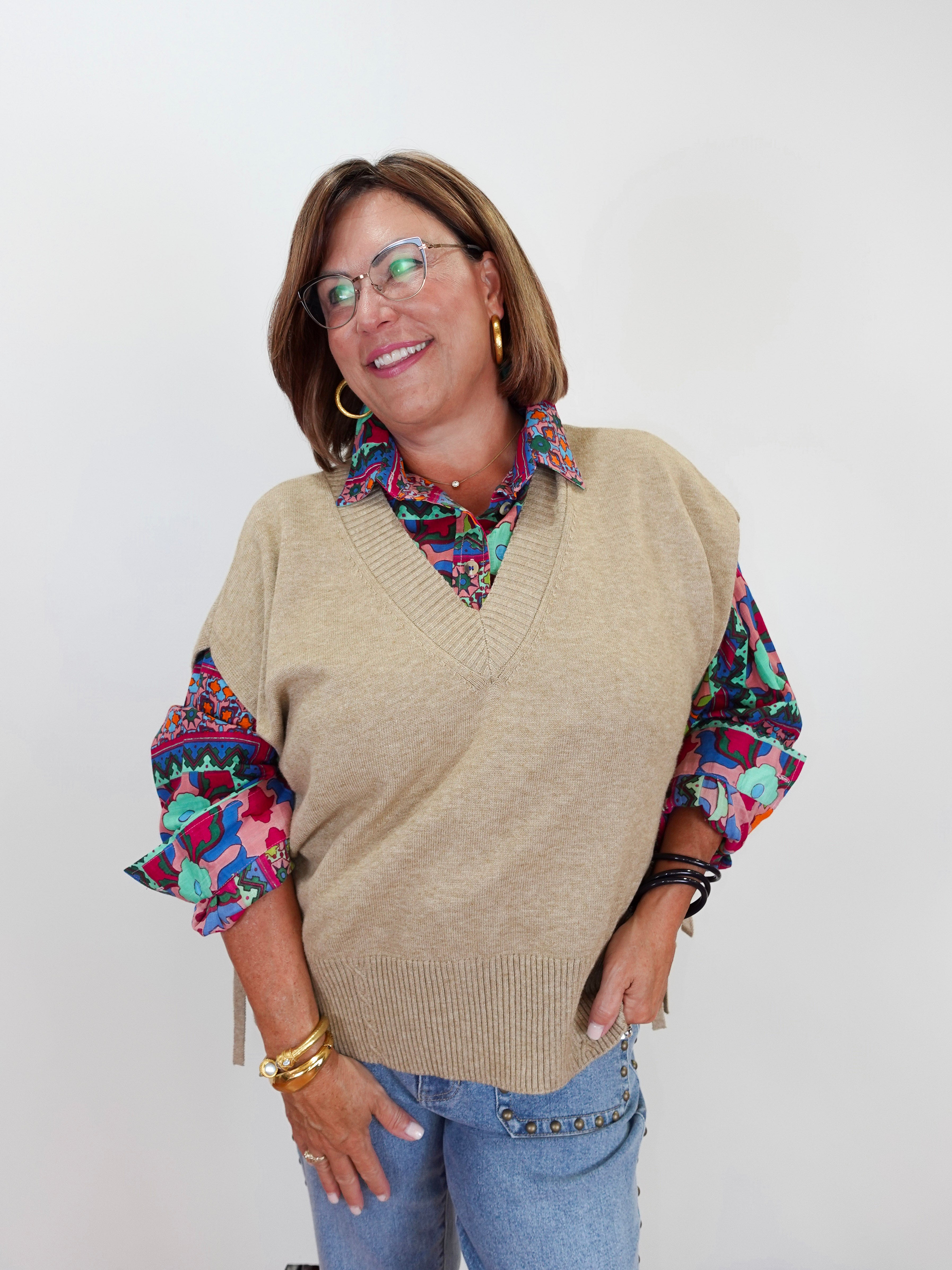 A woman wearing a beige knit V-neck sweater vest over a colorful patterned collared shirt and blue jeans, standing against a plain light gray background.