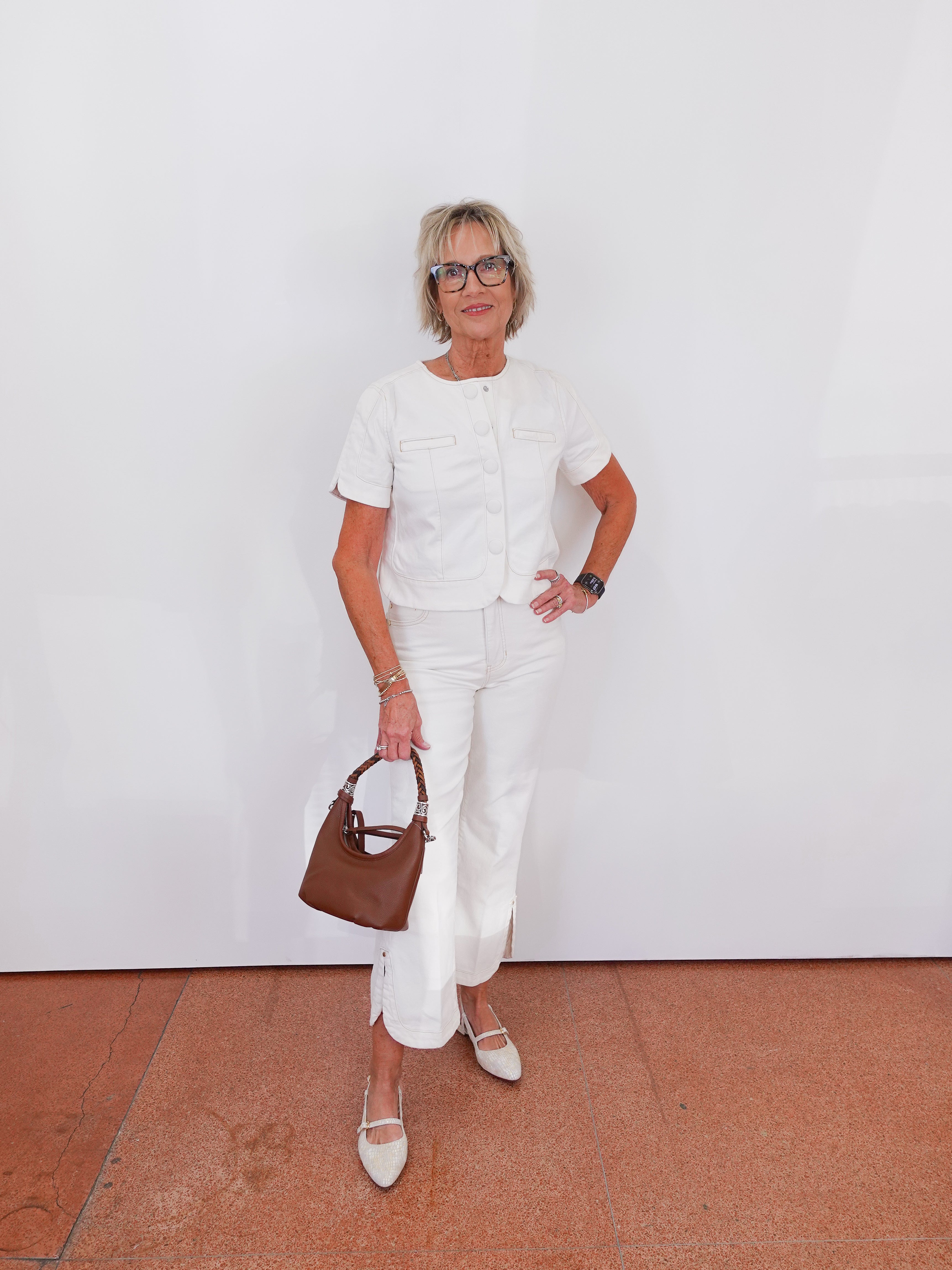Woman in a white outfit holding a brown handbag against a white wall.