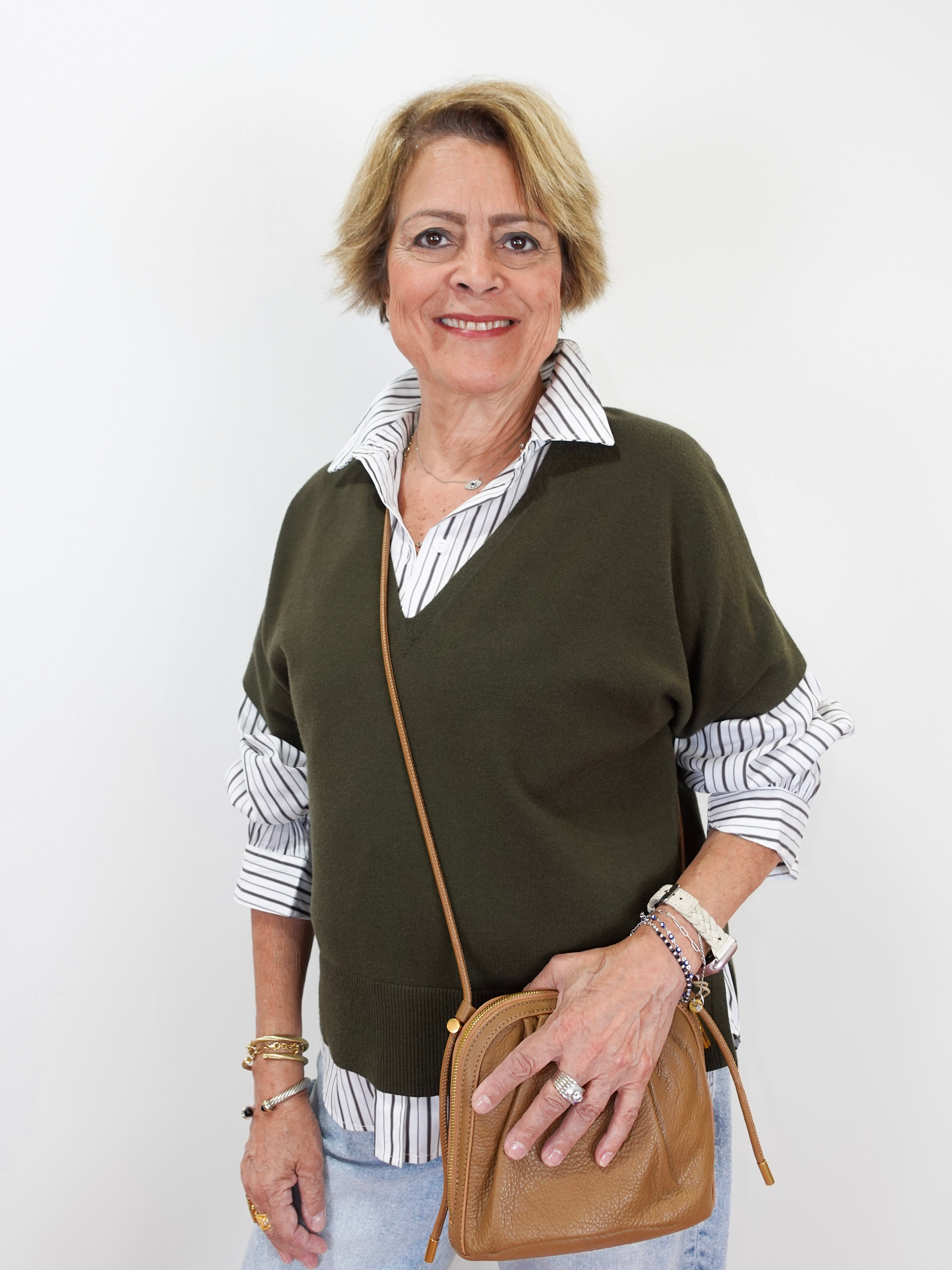 An older woman with short blonde hair is shown from the waist up, wearing a dark olive green knit sweater vest over a white and navy striped button-down shirt, paired with light blue jeans, a tan leather crossbody bag, and various bracelets.