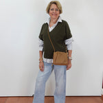 A woman stands against a plain white wall wearing a dark olive green knit sweater vest over a white and navy striped button-down shirt, paired with light-wash wide-leg jeans, black sandals, and a tan crossbody bag.