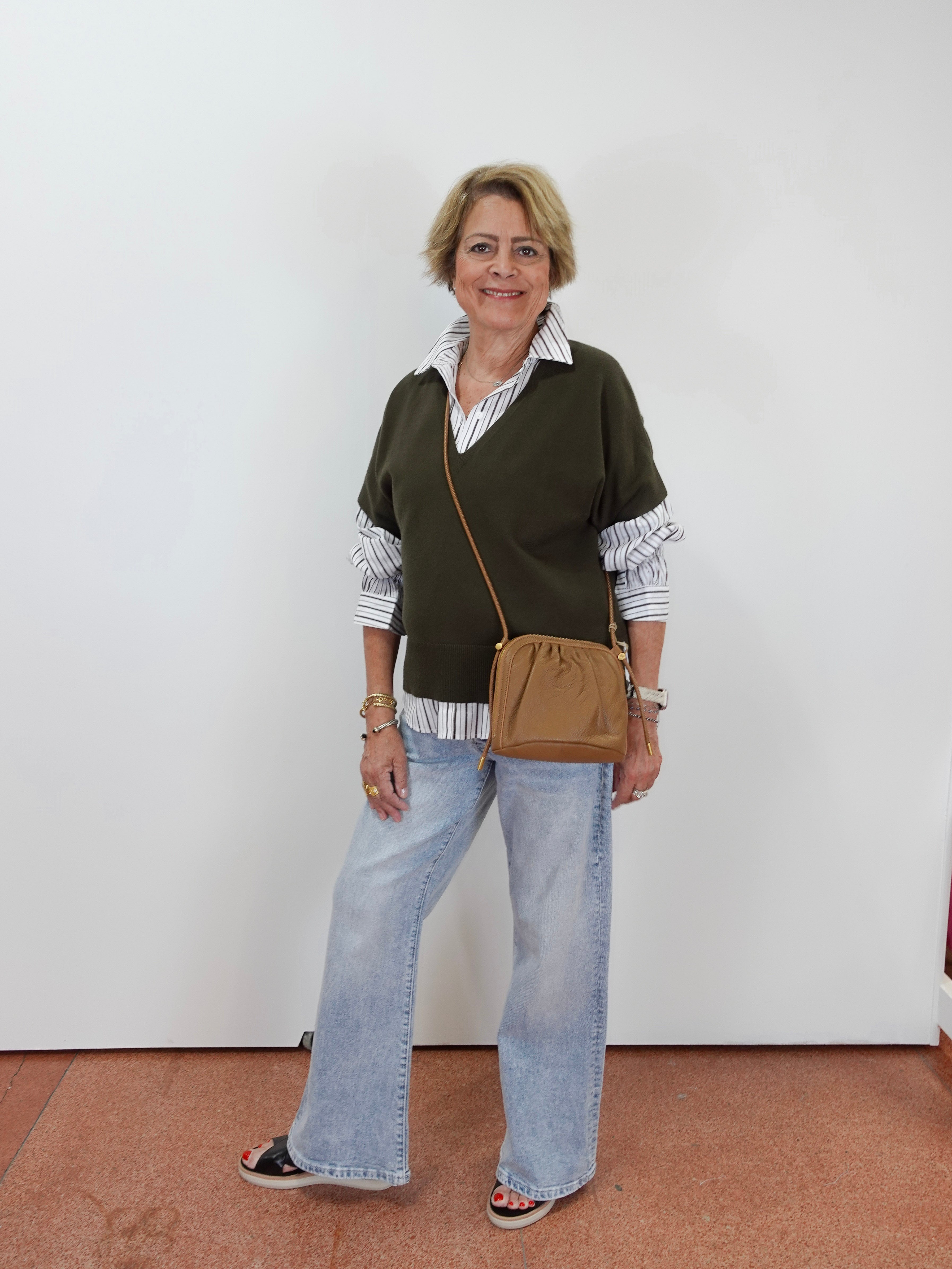 A woman stands against a plain white wall wearing a dark olive green knit sweater vest over a white and navy striped button-down shirt, paired with light-wash wide-leg jeans, black sandals, and a tan crossbody bag.