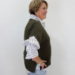 A woman shown in profile wearing a dark olive green knit sweater layered over a white button-down shirt with thin black stripes, paired with light blue jeans, against a plain white background.