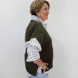 A woman shown in profile wearing a dark olive green knit sweater layered over a white button-down shirt with thin black stripes, paired with light blue jeans, against a plain white background.