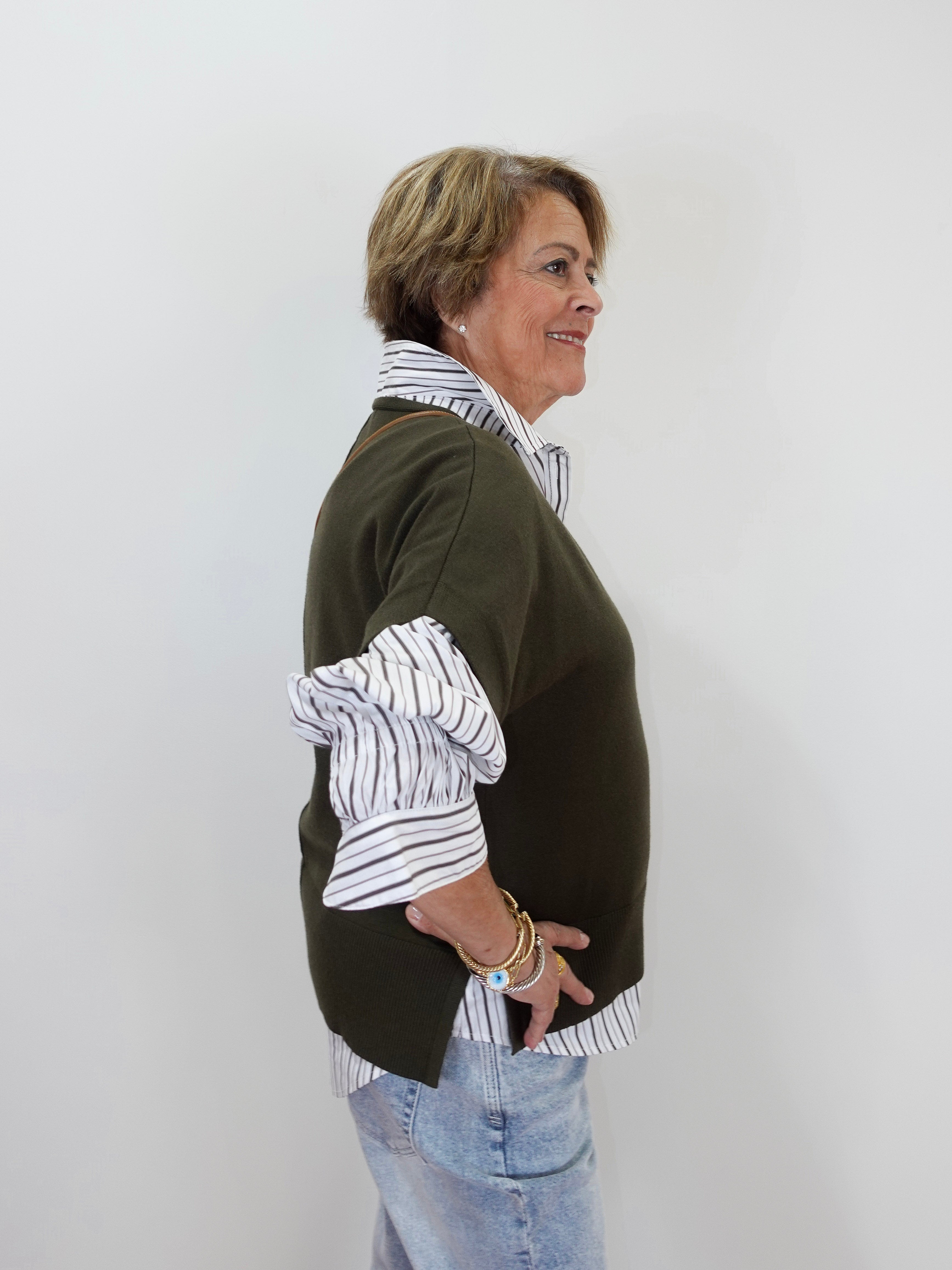 A woman shown in profile wearing a dark olive green knit sweater layered over a white button-down shirt with thin black stripes, paired with light blue jeans, against a plain white background.
