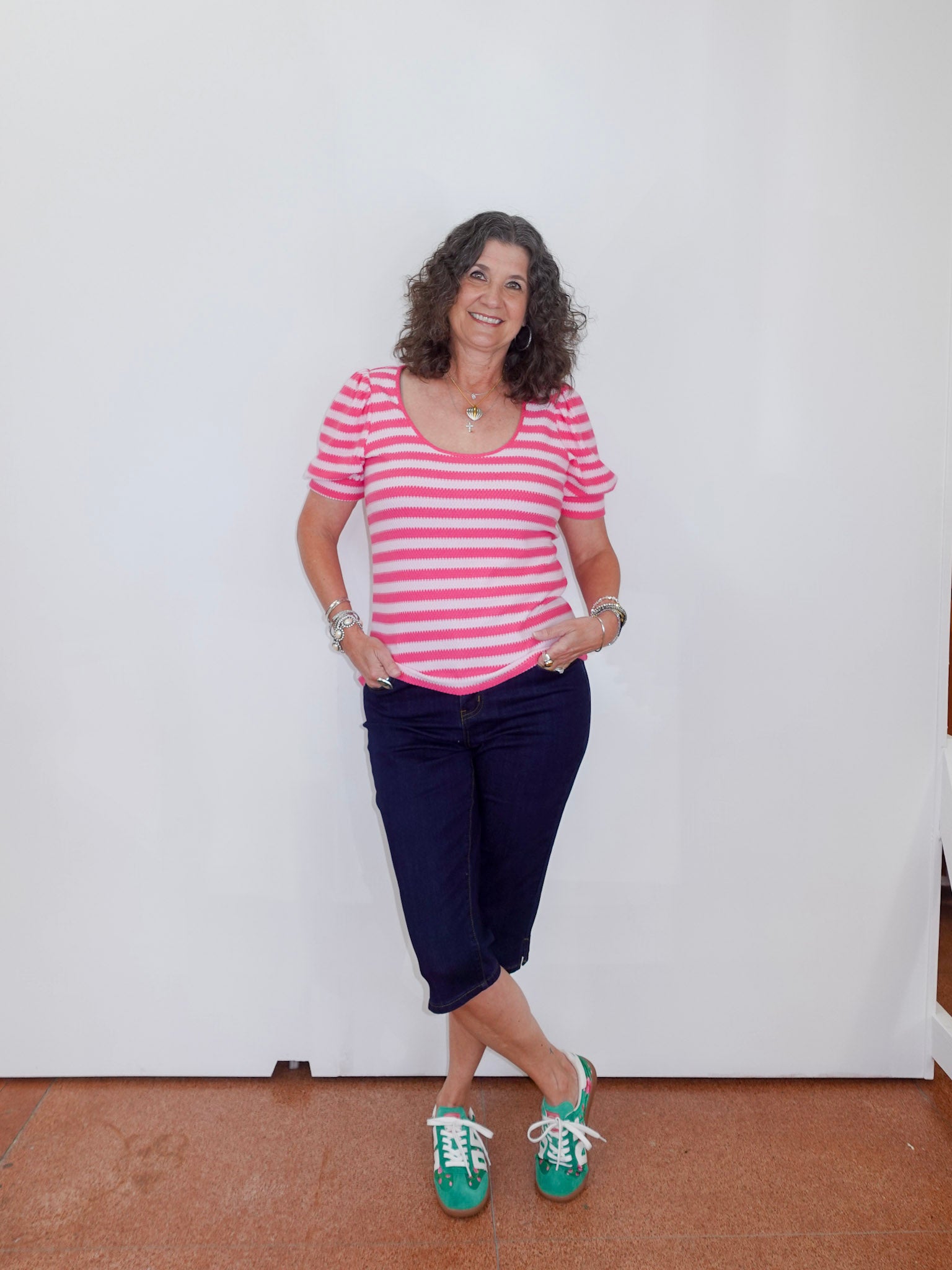 Woman wearing a pink and white striped shirt, navy blue pants, and green sneakers against a white wall.