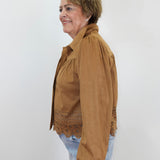 A woman shown in profile wearing a tan long-sleeve button-up shirt jacket with eyelet lace trim at the hem, paired with light blue jeans, against a plain white background.