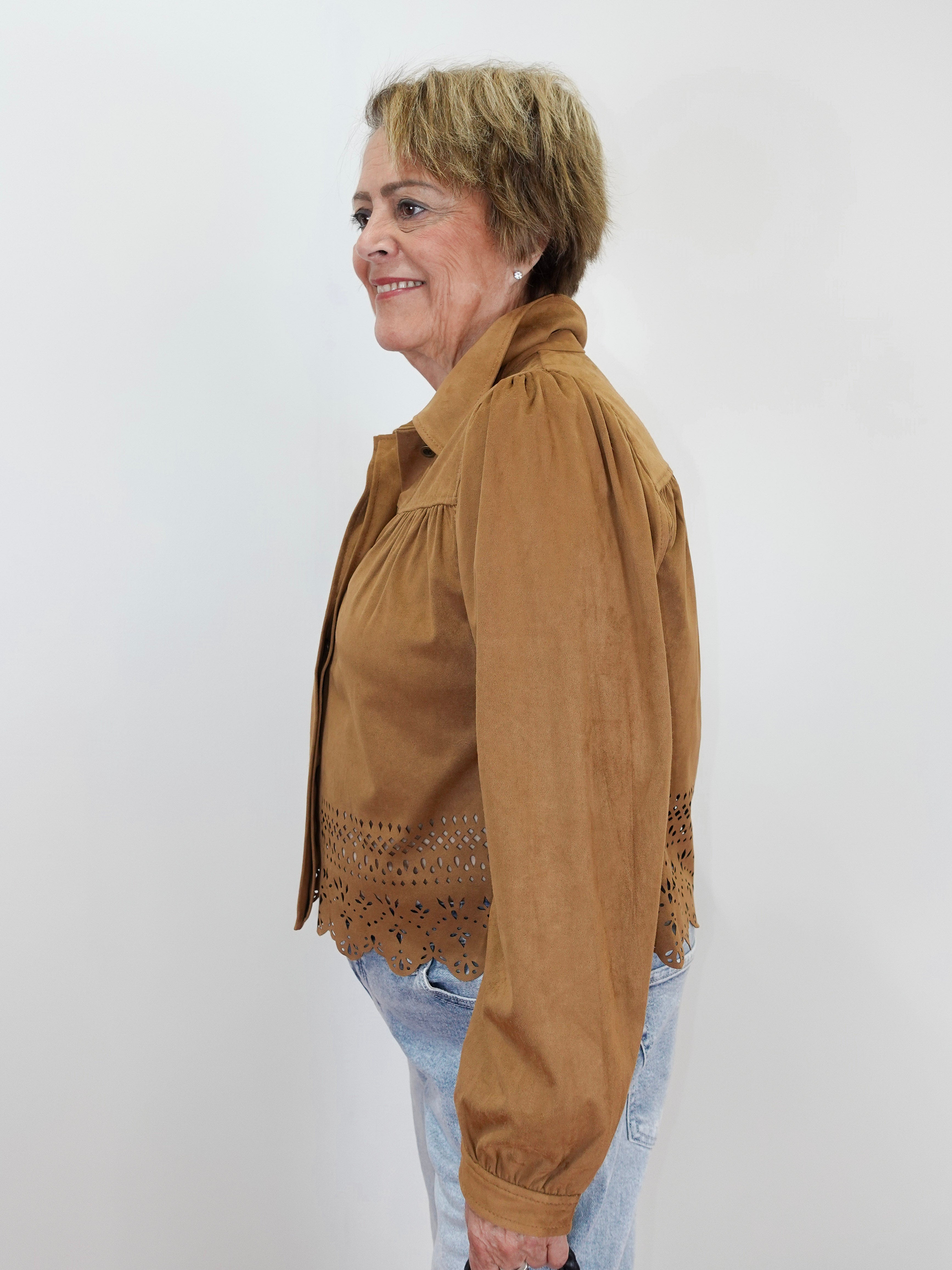 A woman shown in profile wearing a tan long-sleeve button-up shirt jacket with eyelet lace trim at the hem, paired with light blue jeans, against a plain white background.