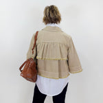 Person wearing a beige jacket with fringe details, holding a brown bag against a white background