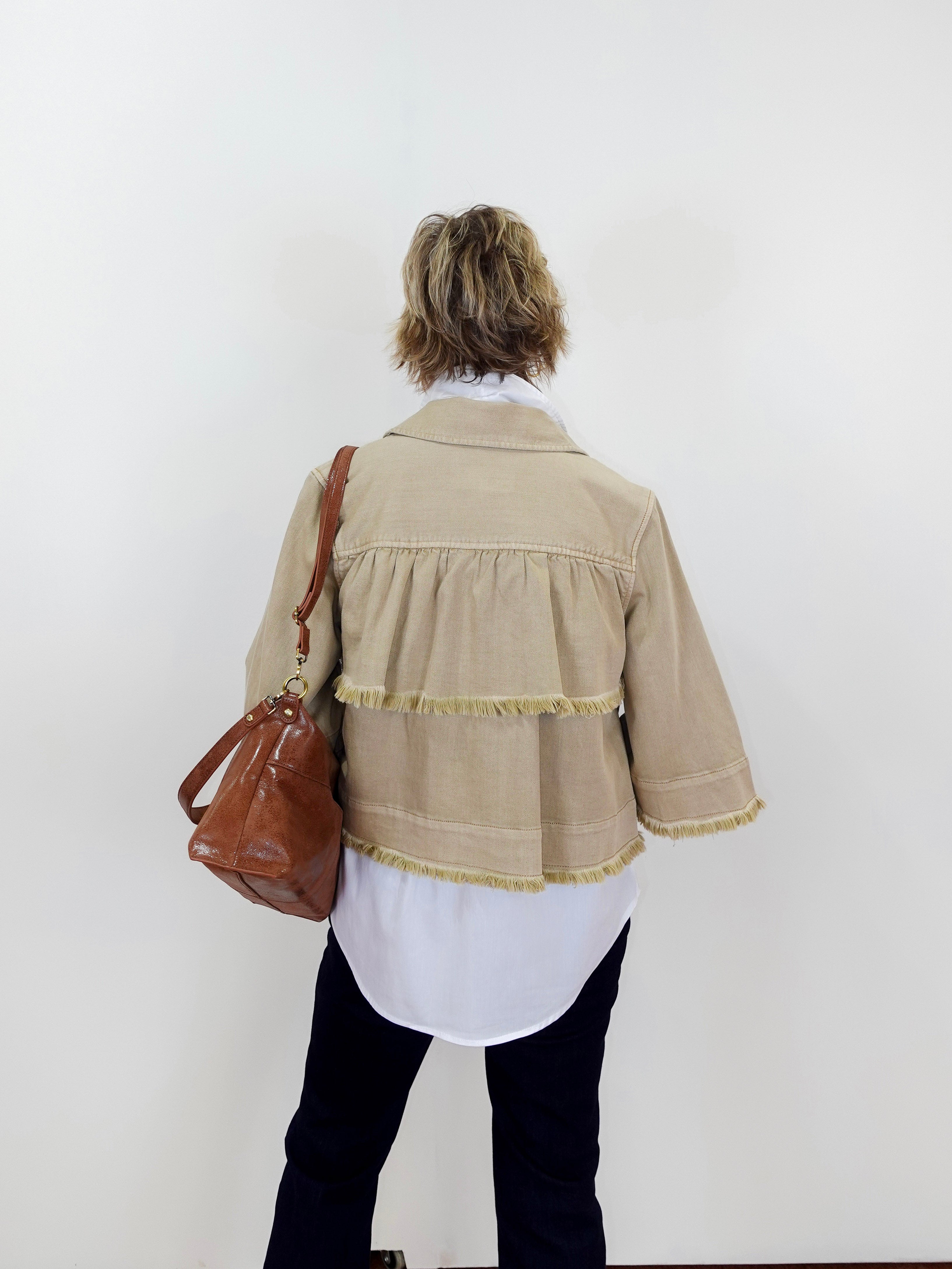 Person wearing a beige jacket with fringe details, holding a brown bag against a white background