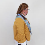 A woman wearing a mustard yellow jacket, a white shirt, black pants, and a blue patterned scarf, standing against a plain white background.