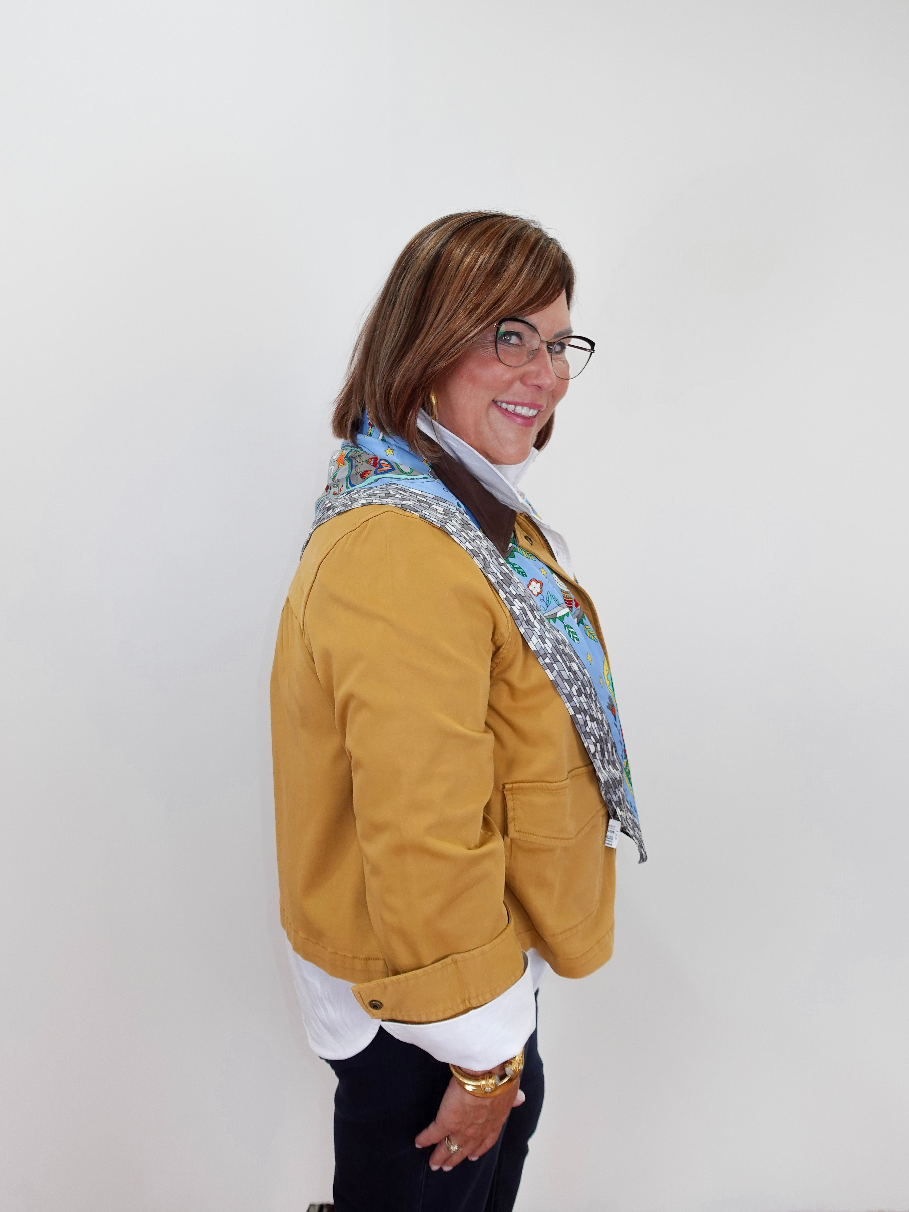 A woman wearing a mustard yellow jacket, a white shirt, black pants, and a blue patterned scarf, standing against a plain white background.